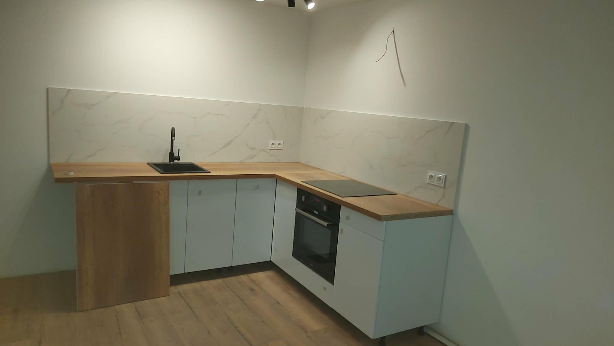 Nowoczesna kuchnia z białymi szafkami, drewnianym blatem i marmurowym panelem ściennym. Czarny zlew i płyta indukcyjna. Minimalistyczny design, jasne wnętrze.