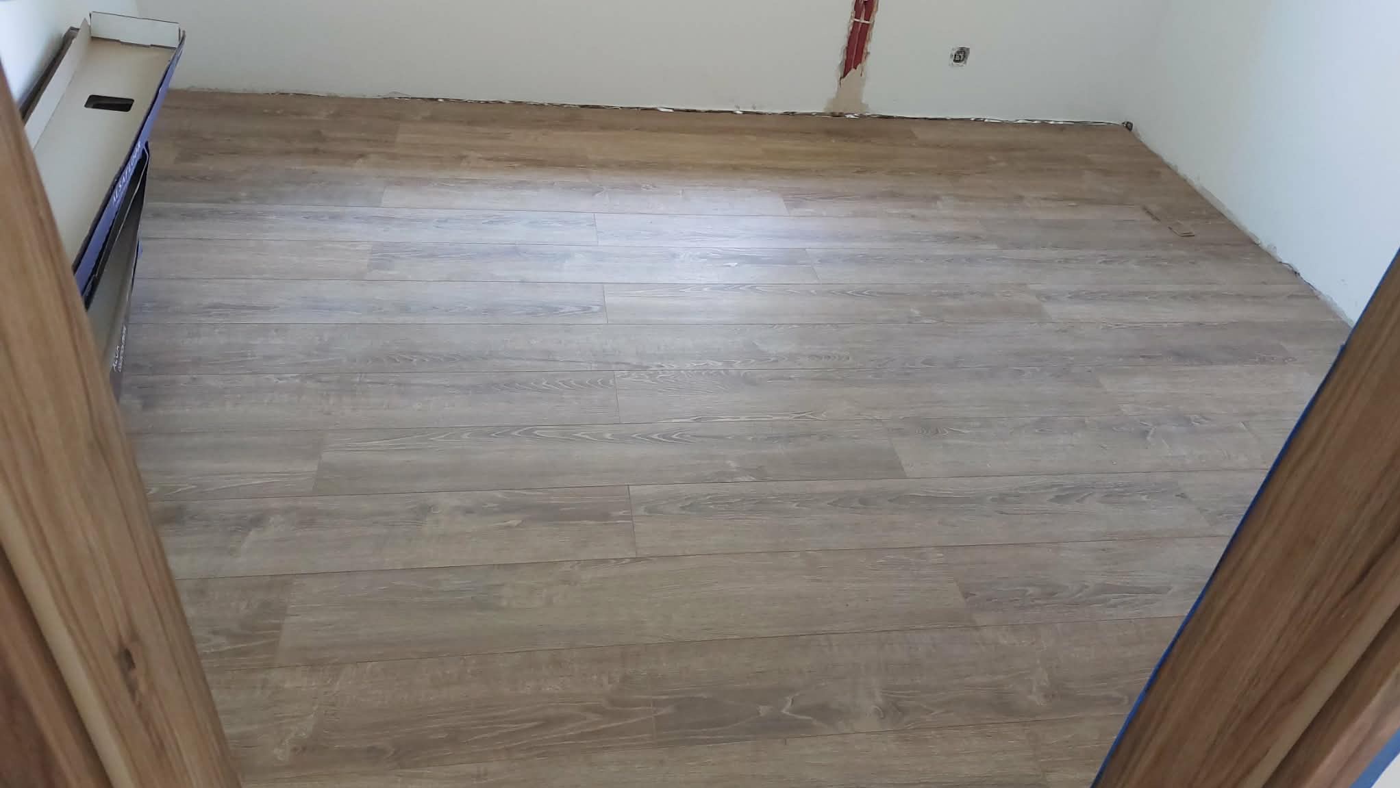 Ułożony parkiet w jasnym odcieniu drewna, widok z góry, z fragmentem kartonu po lewej. Ściany białe, w tle widoczne gniazdko elektryczne i instalacja.