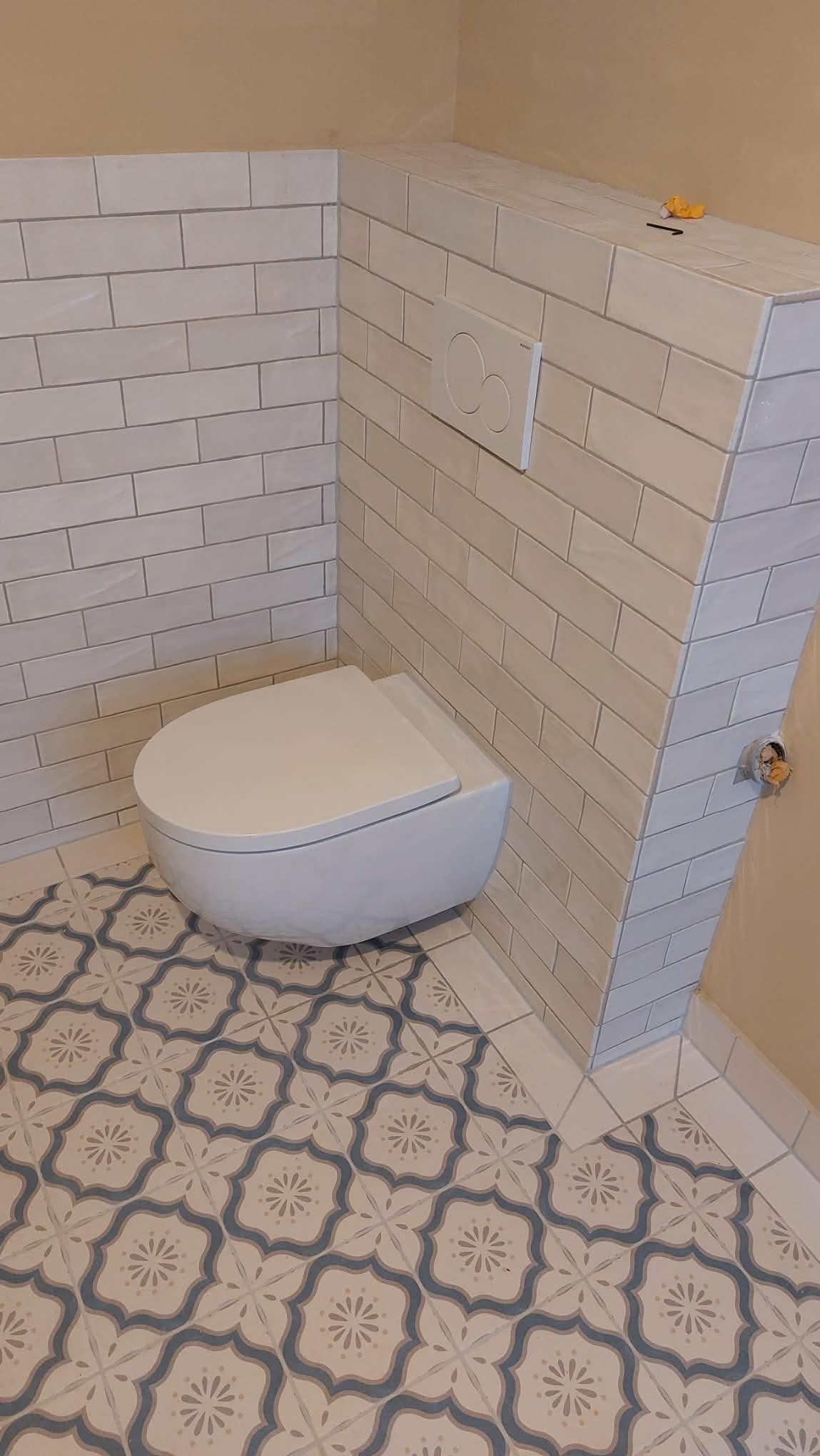 Nowoczesna toaleta podwieszana w wykończonej łazience. Białe płytki na ścianach i wzorzysta podłoga tworzą spójną całość. Widoczny przycisk spłuczki i fragment instalacji.