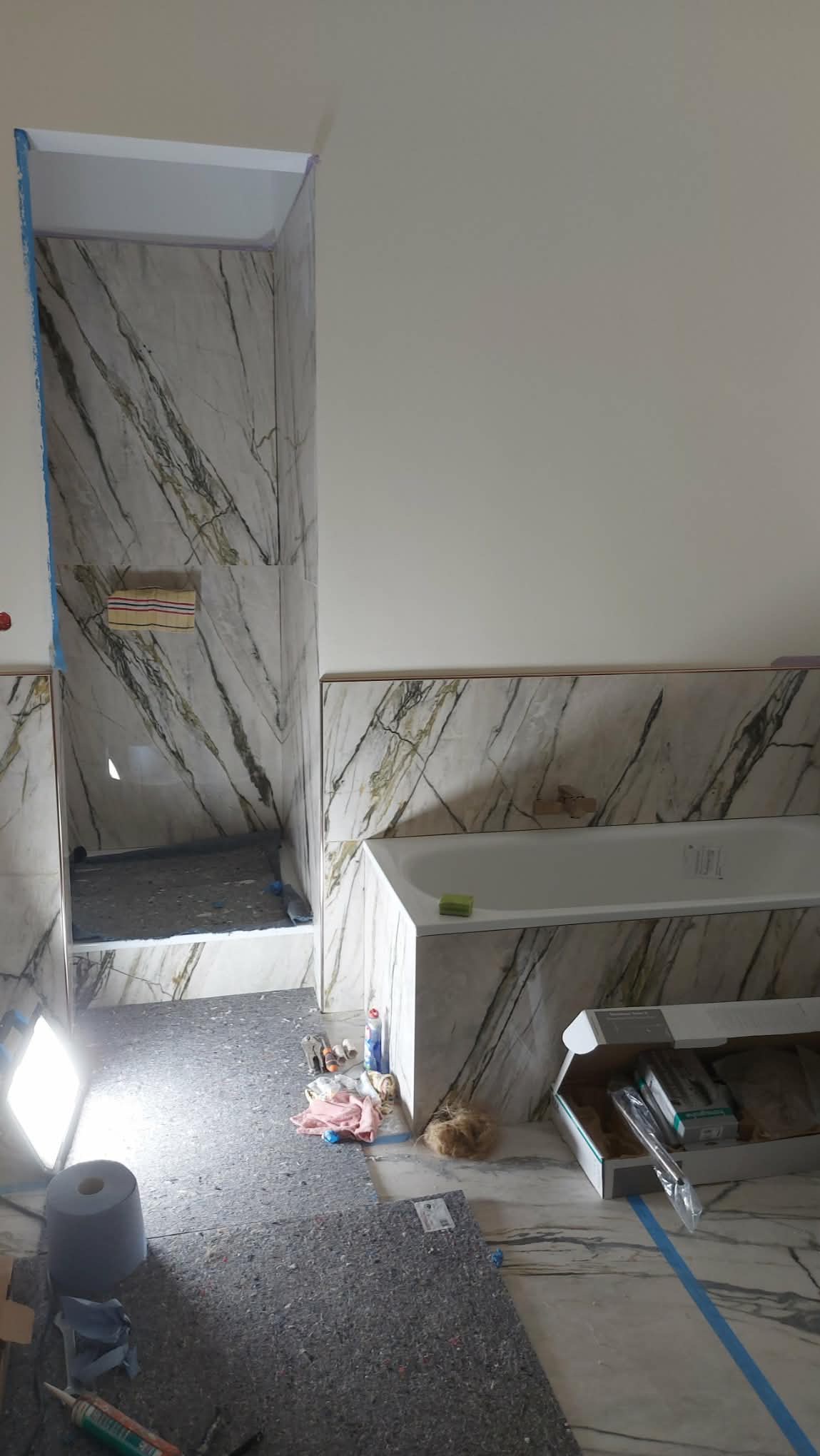 Wykończenie łazienki w trakcie: marmurowe płytki z zielonymi żyłami na ścianach w strefie prysznica i wanny. Widoczne narzędzia i materiały budowlane.