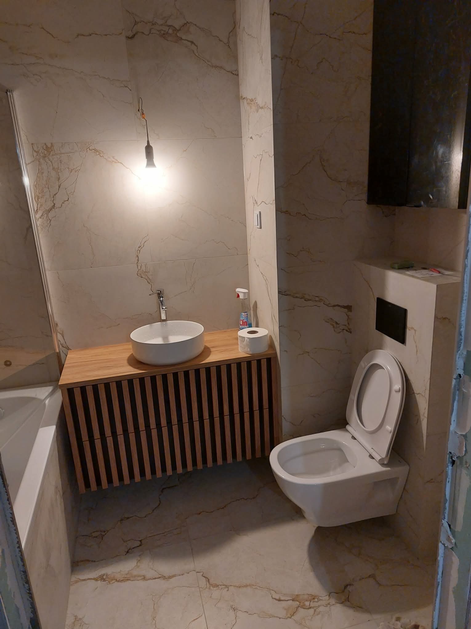 Nowoczesna łazienka z marmurowymi płytkami, wiszącą toaletą i szafką pod umywalkę z drewnianymi lamelami. Widoczna bateria i akcesoria łazienkowe.