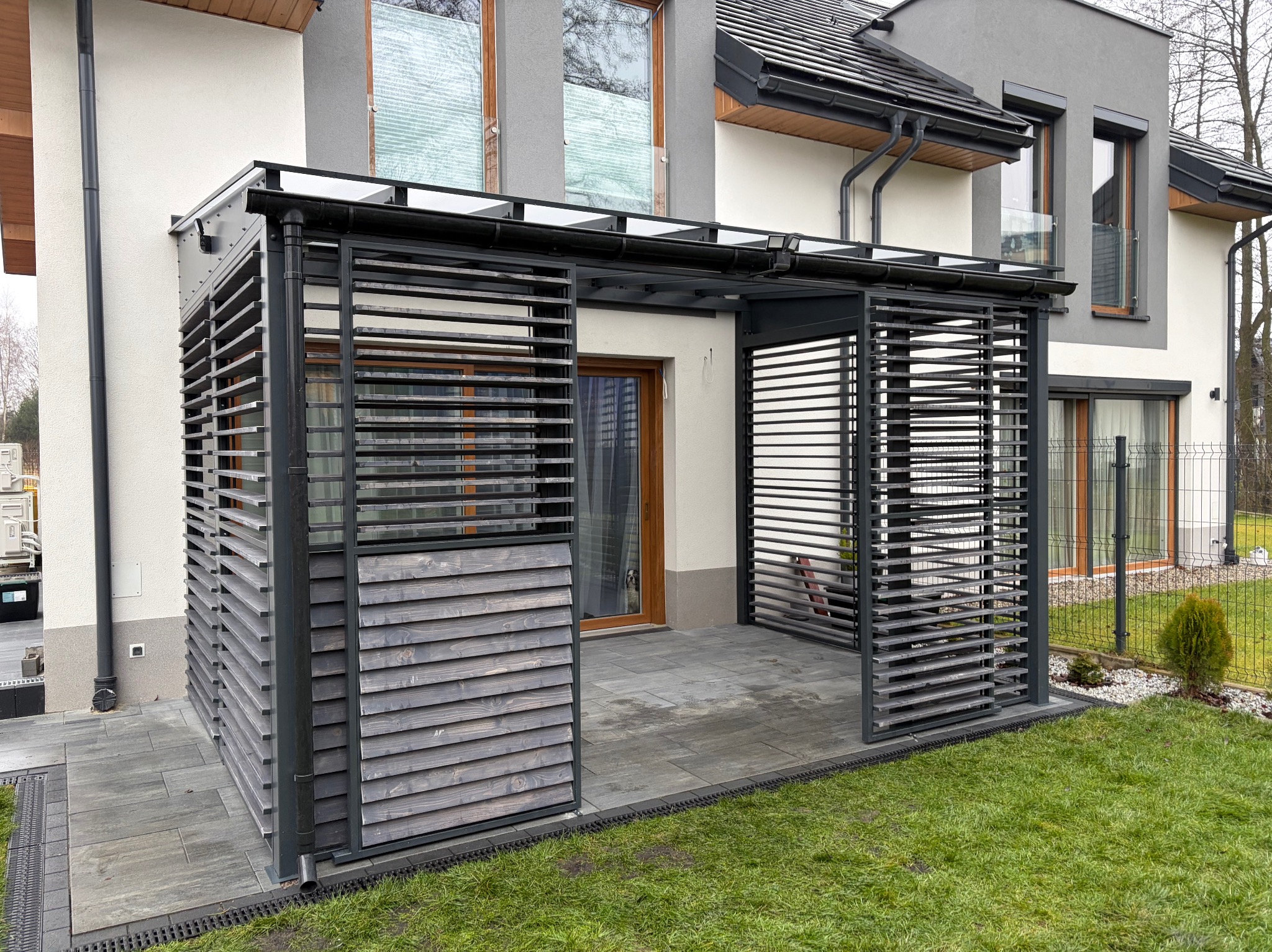 Nowoczesna pergola z przesuwnymi, drewnianymi lamelami w kolorze grafitowym, zadaszona poliwęglanem, na tle domu z szarą elewacją i zielonym trawnikiem.