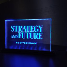 Anna Skowrońska ARTCOP - Podświetlana, akrylowa tabliczka z napisem 'Strategy and Future' w kolorze niebieskim, umieszczona na ciemnym tle. Nowoczesny design, wyraźne liternictwo.