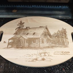 Anna Skowrońska ARTCOP - Deska z jasnego drewna z wygrawerowanym laserowo rysunkiem chaty i drzew, wykonana techniką CNC. Widoczne logo firmy Artcop.