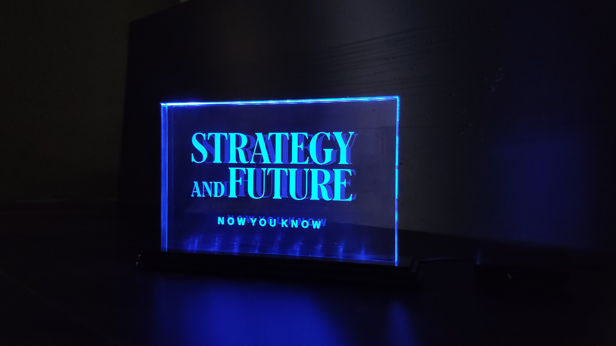 Podświetlana, akrylowa tabliczka z napisem 'Strategy and Future' w kolorze niebieskim, umieszczona na ciemnym tle. Nowoczesny design, wyraźne liternictwo.
