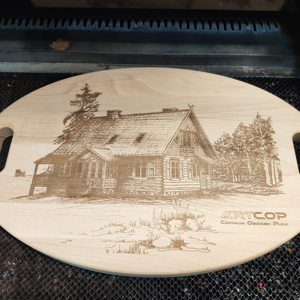 Deska z jasnego drewna z wygrawerowanym laserowo rysunkiem chaty i drzew, wykonana techniką CNC. Widoczne logo firmy Artcop.