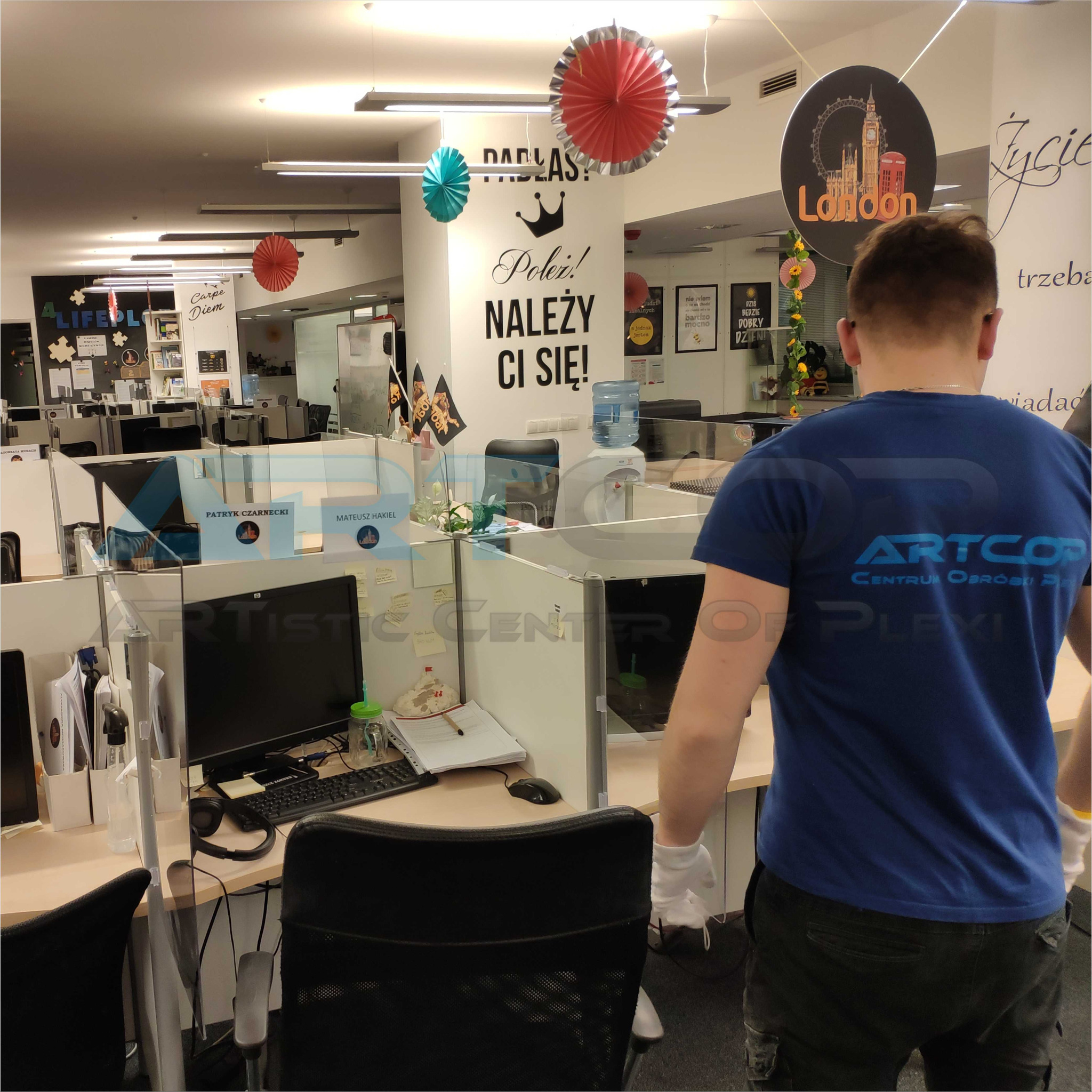 Pracownik w niebieskiej koszulce z logo firmy sprzątającej biuro z przegrodami z pleksi. Widoczne biurka z komputerami i dekoracje biurowe.