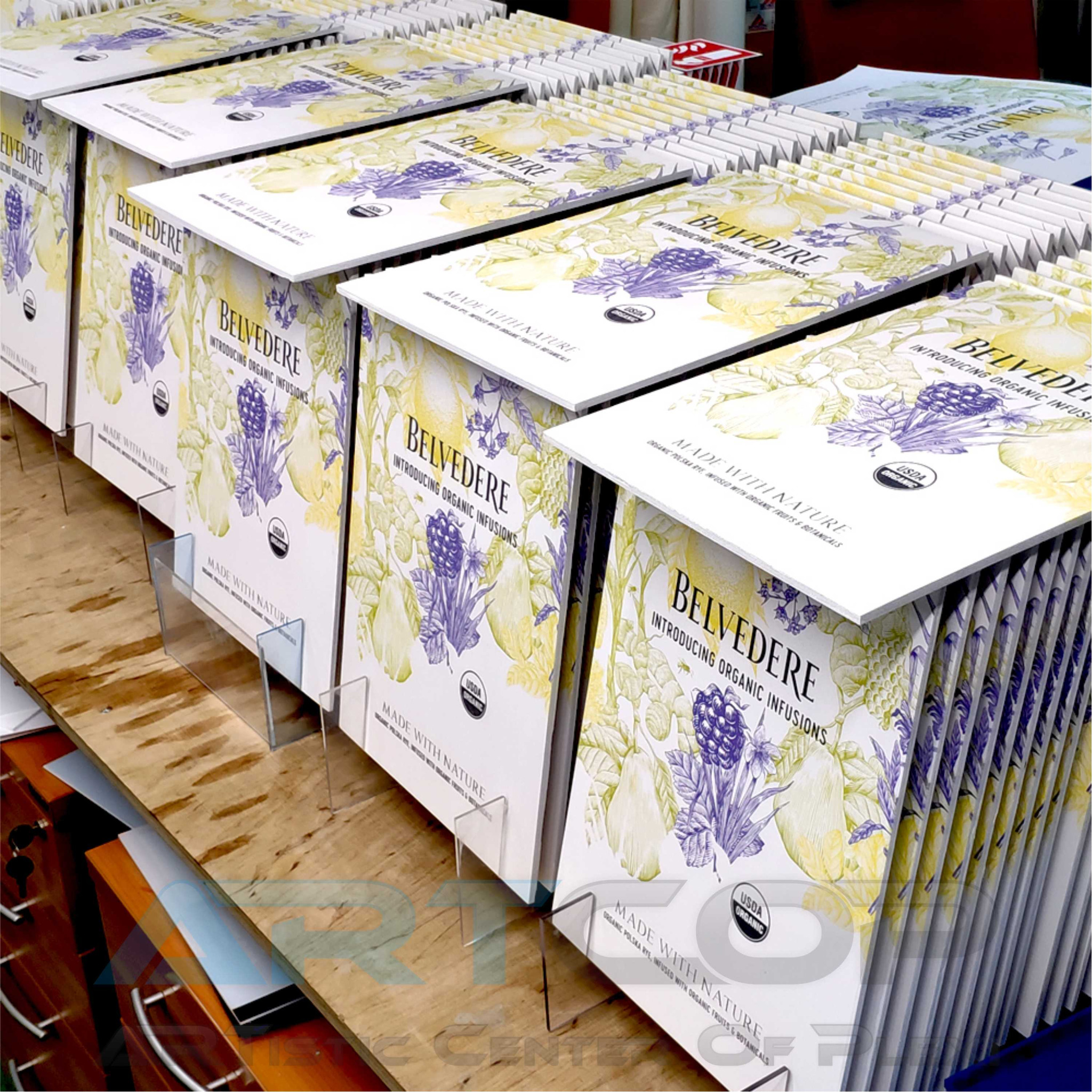 Stojaki z ulotkami Belvedere Organic Infusions, ustawione rzędami na drewnianym blacie. Grafika z motywami roślinnymi w odcieniach fioletu i zieleni. Widoczne logo USDA Organic.