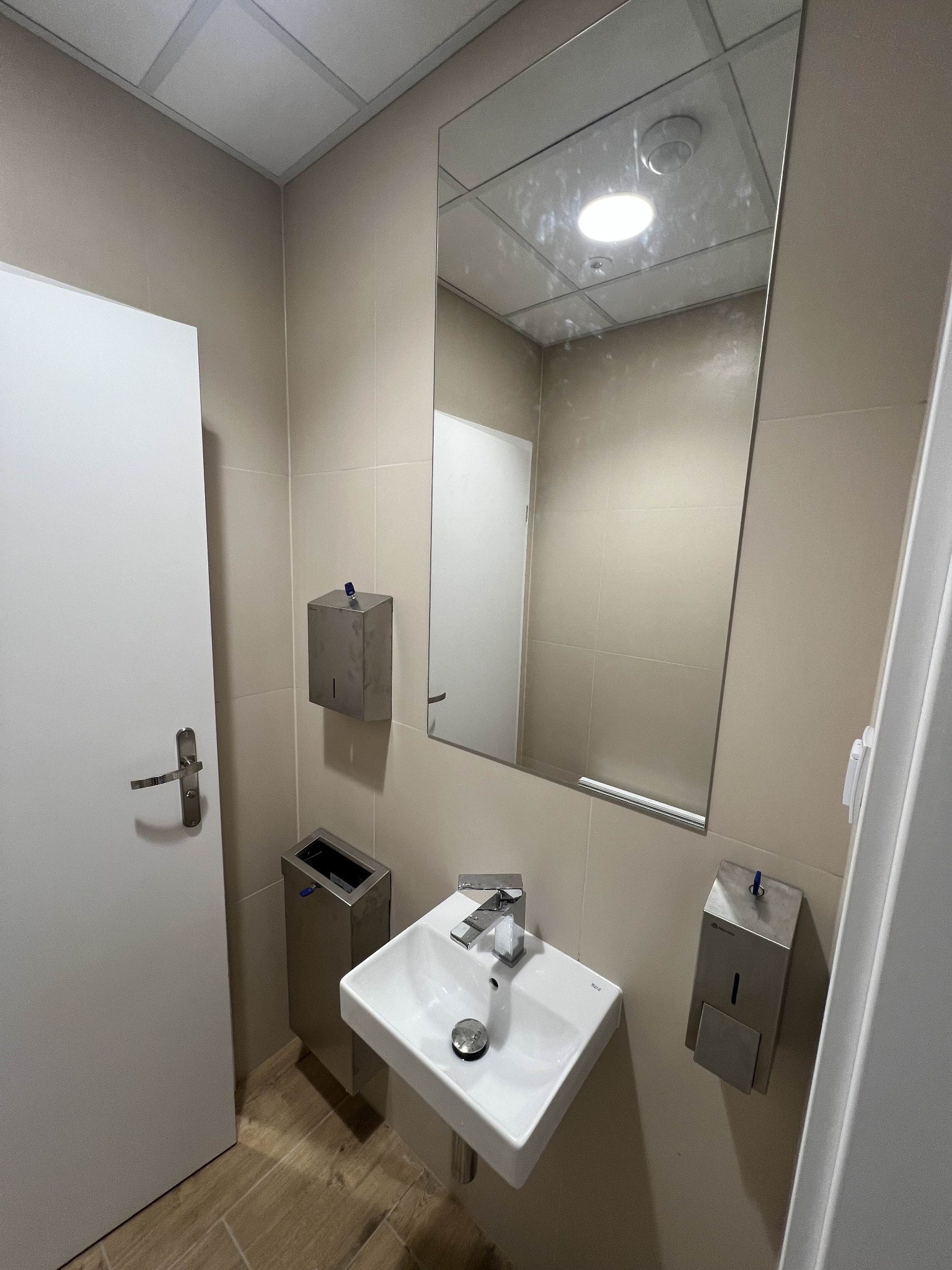 Jasna łazienka z umywalką, lustrem i dozownikiem mydła. Beżowe płytki na ścianach i drewnopodobna podłoga. Widoczna biała ściana i drzwi.