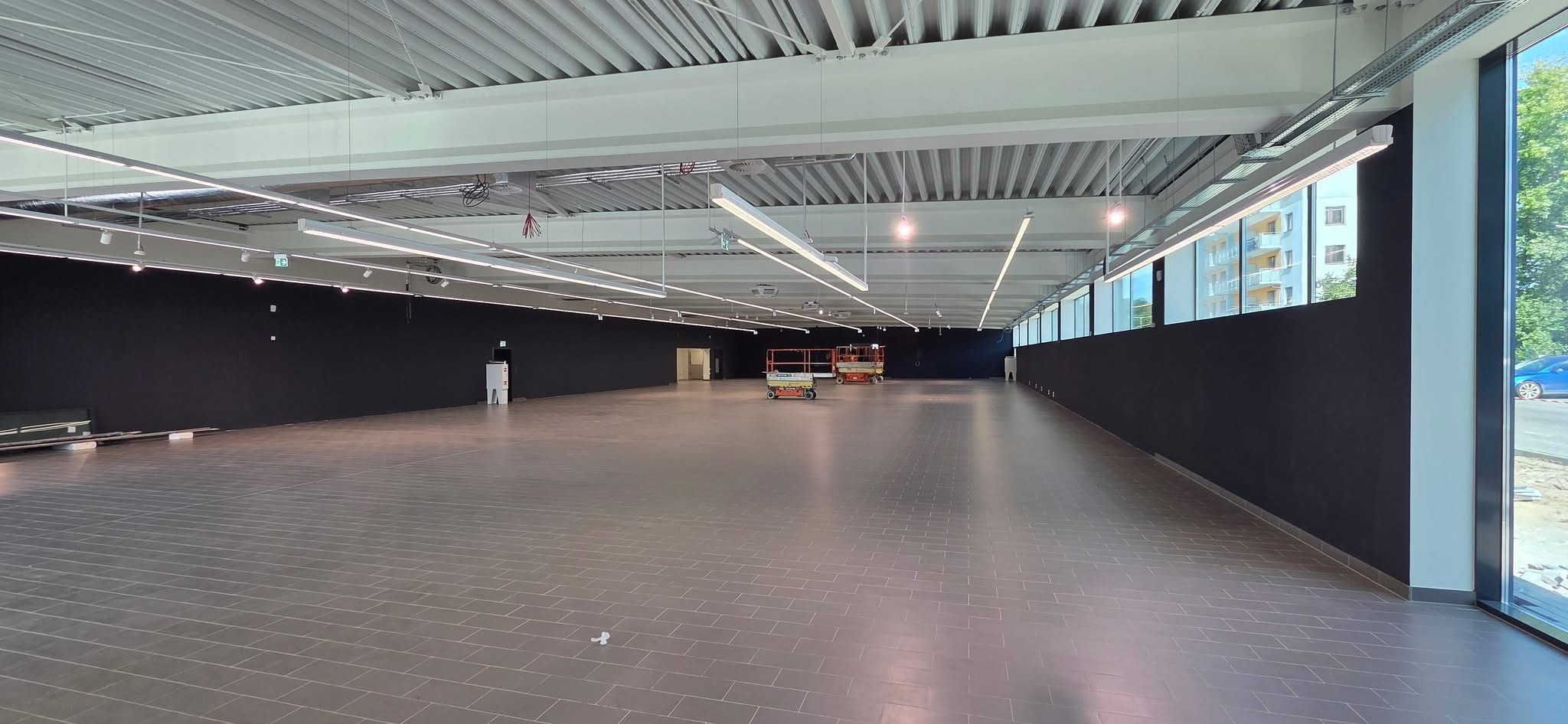 Przestronne, puste wnętrze biurowe z szarą podłogą, czarnymi ścianami i białym sufitem z oświetleniem. Dwie podnośniki koszowe w oddali.