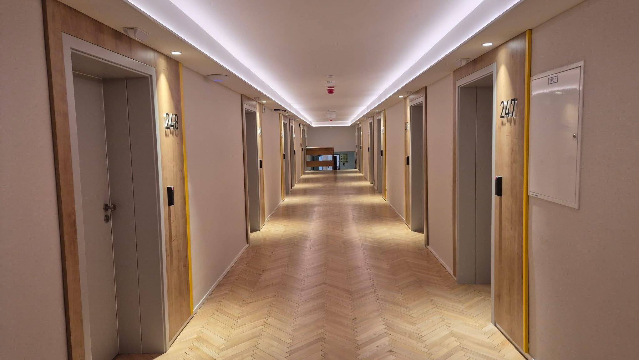 Długi korytarz z drewnianą podłogą w jodełkę, oświetlony ledami. Drzwi z numerami i drewnianymi obramowaniami po obu stronach. Widok w głąb korytarza.