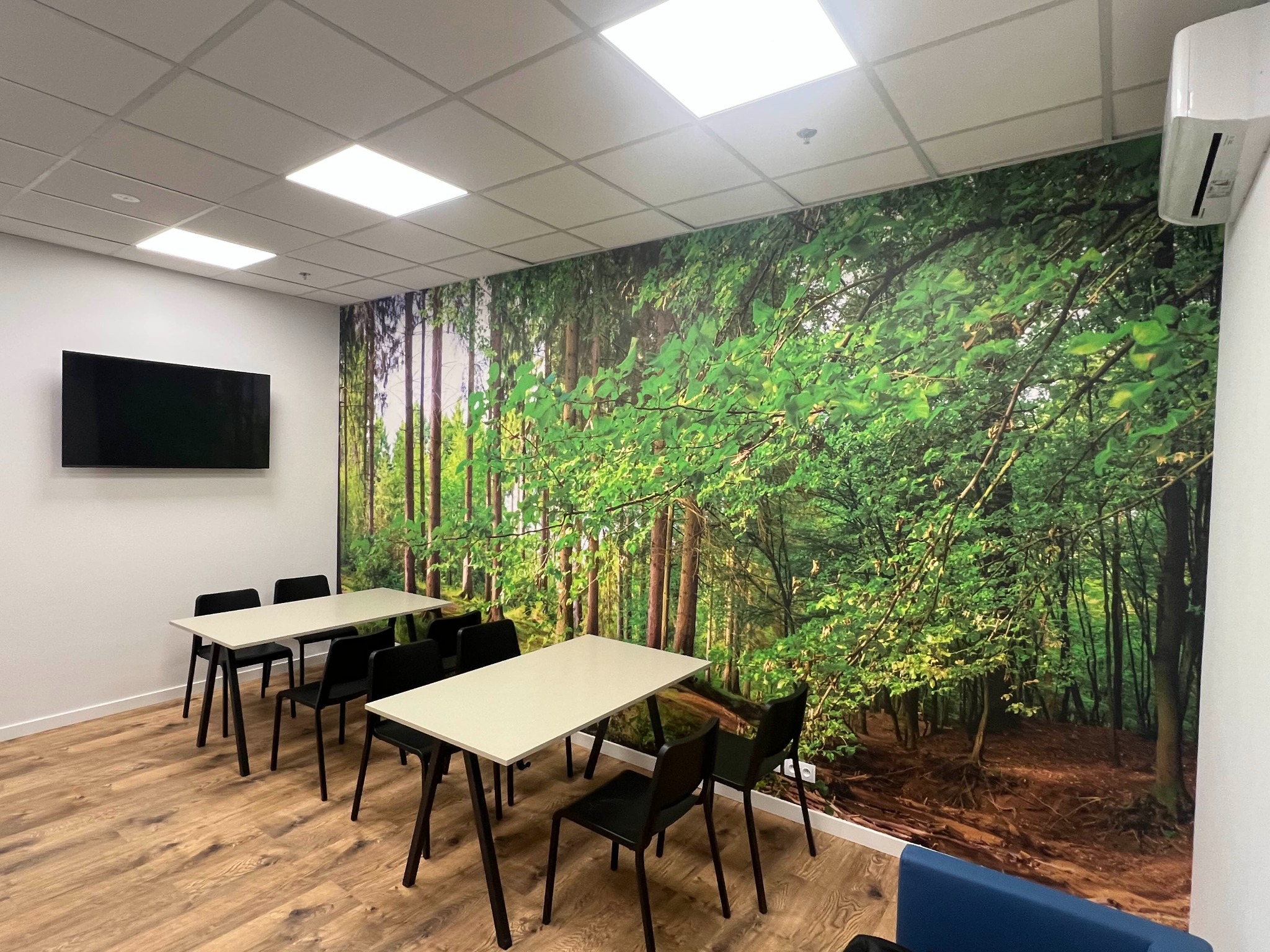 Nowoczesne biuro po remoncie: fototapeta z motywem leśnym na ścianie, drewniana podłoga, białe stoły i czarne krzesła. Klimatyczne oświetlenie sufitowe.