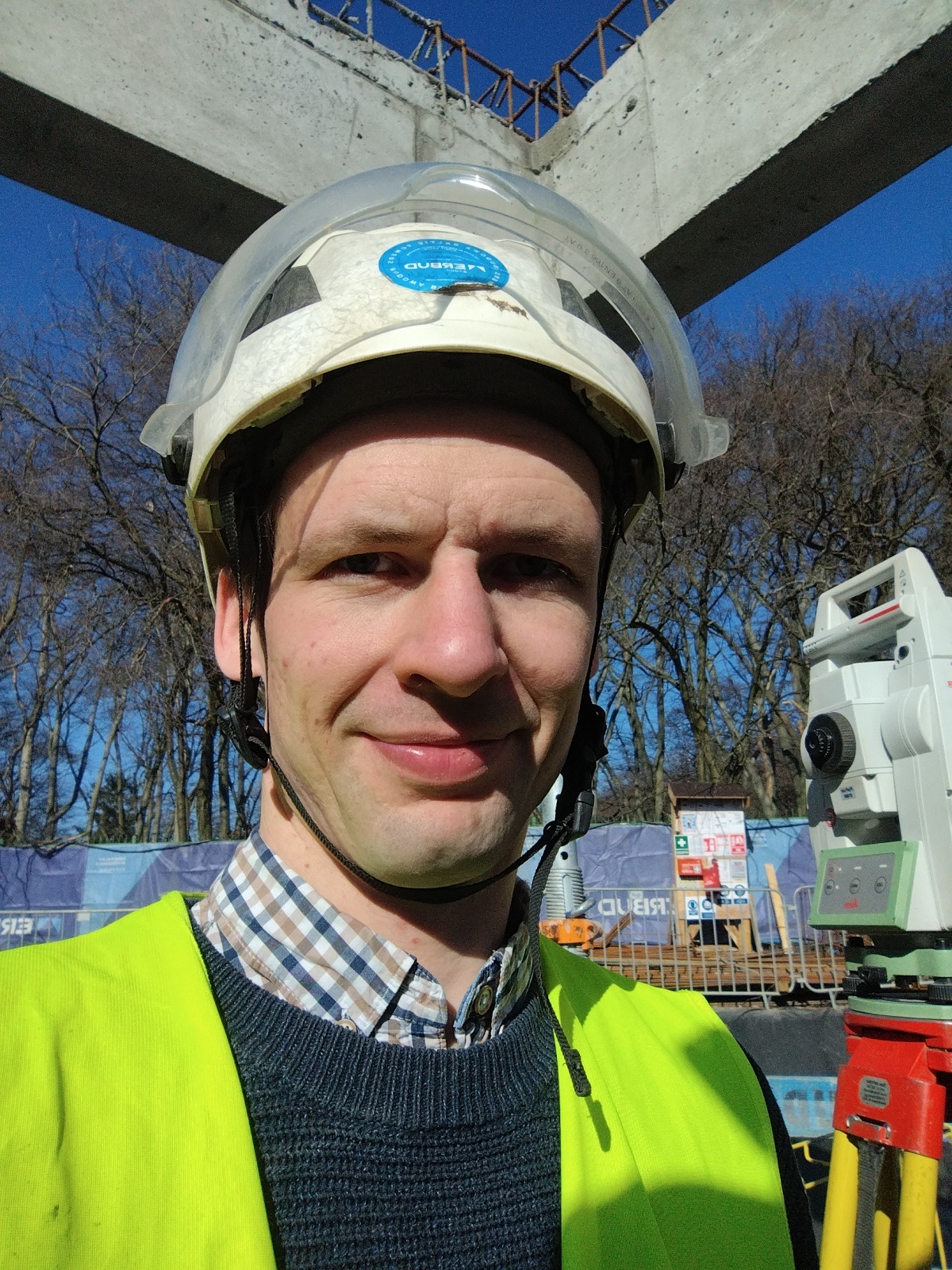 Uśmiechnięty geodeta w kasku i kamizelce odblaskowej, z niwelatorem w tle, na tle konstrukcji betonowej i drzew. Słoneczny dzień.