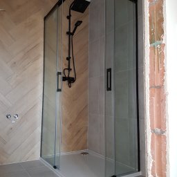 Daniel Pietrzyk - Nowoczesna kabina prysznicowa z czarnymi profilami i deszczownicą, na tle ściany wyłożonej płytkami imitującymi drewno. Widoczny fragment surowej ściany.