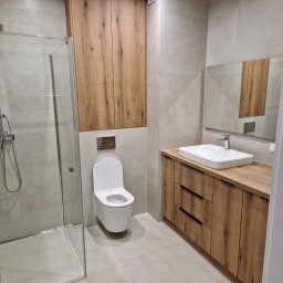 Daniel Pietrzyk - Nowoczesna łazienka z kabiną prysznicową, toaletą i umywalką na szafce z drewnianym wykończeniem. Jasne płytki na ścianach i podłodze. Minimalistyczny design.