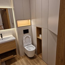 Daniel Pietrzyk - Nowoczesna łazienka po remoncie: podwieszana toaleta, szafki w zabudowie, drewniana podłoga i umywalka z lustrem. Minimalistyczny design z oświetleniem LED.