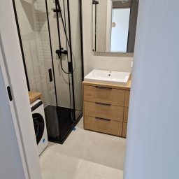 Daniel Pietrzyk - Nowoczesna łazienka po remoncie: kabina prysznicowa typu walk-in z czarną armaturą, drewniana szafka z umywalką i pralka. Minimalistyczny design, jasne płytki.