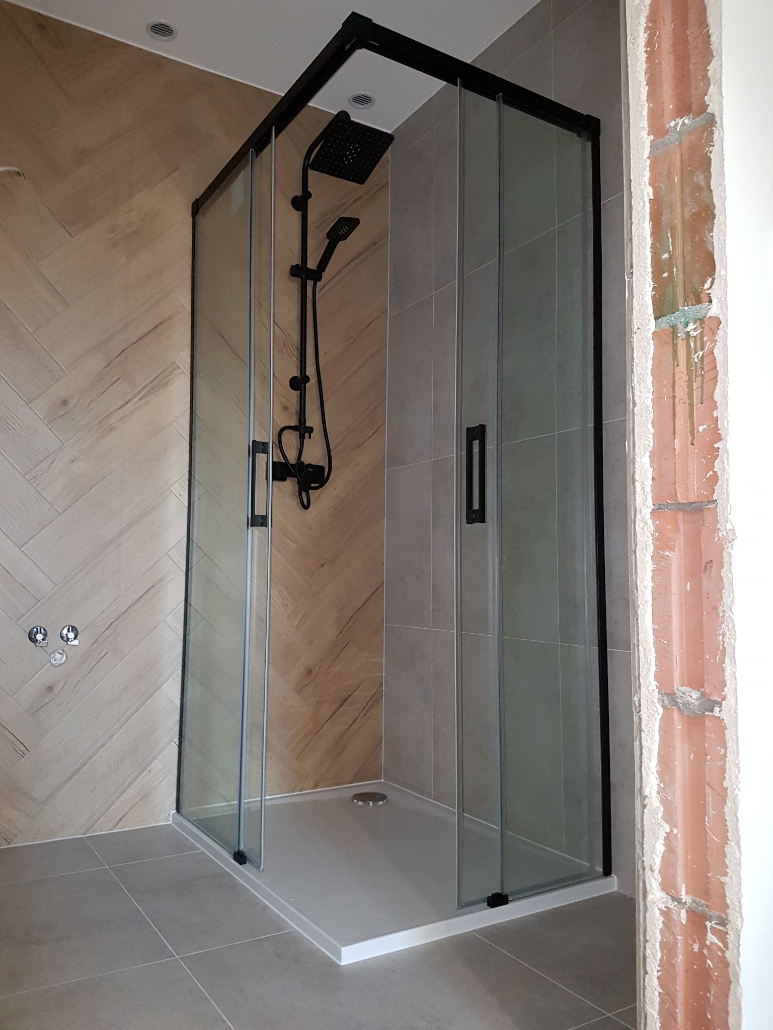 Nowoczesna kabina prysznicowa z czarnymi profilami i deszczownicą, na tle ściany wyłożonej płytkami imitującymi drewno. Widoczny fragment surowej ściany.