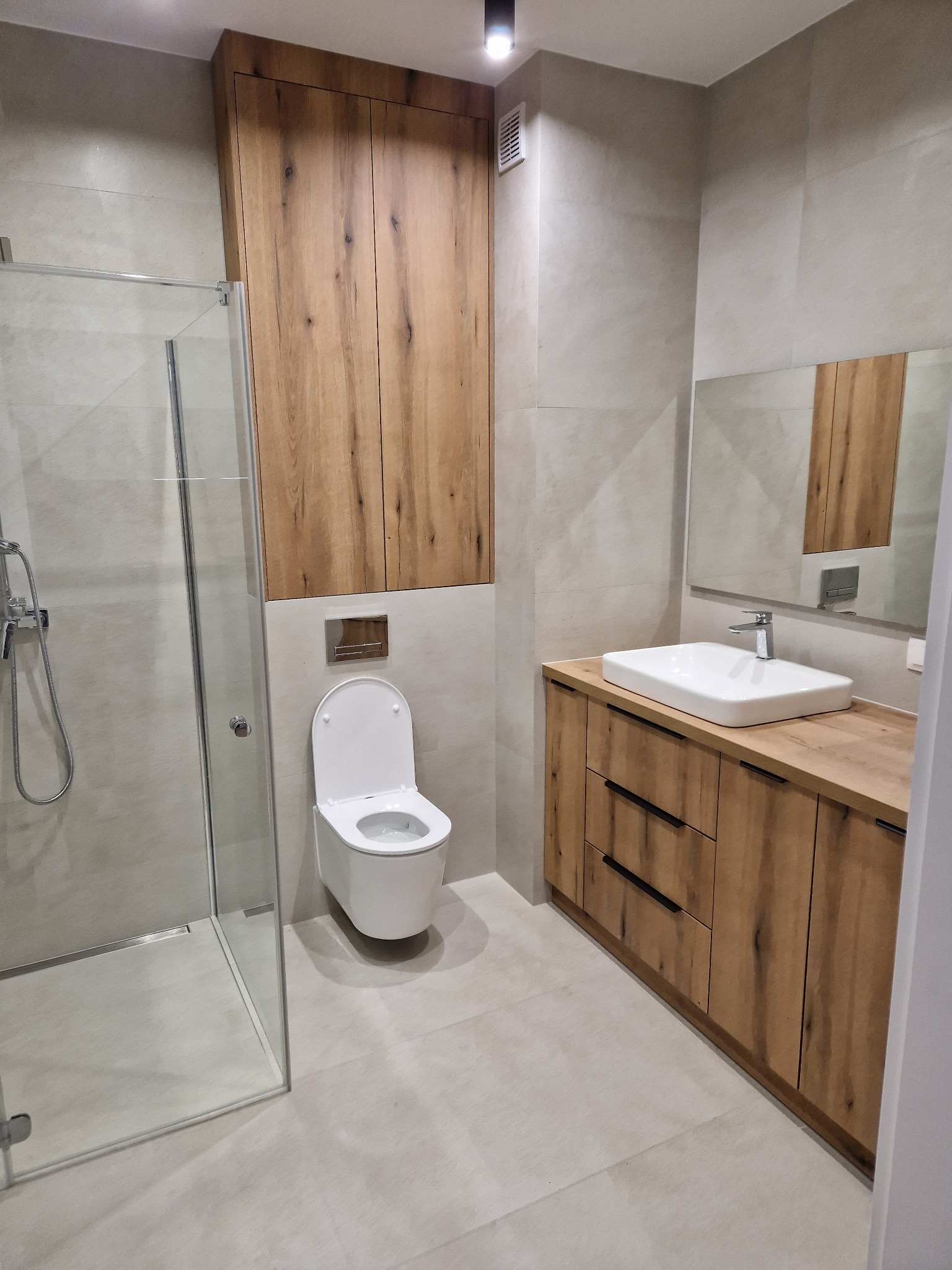 Nowoczesna łazienka z kabiną prysznicową, toaletą i umywalką na szafce z drewnianym wykończeniem. Jasne płytki na ścianach i podłodze. Minimalistyczny design.