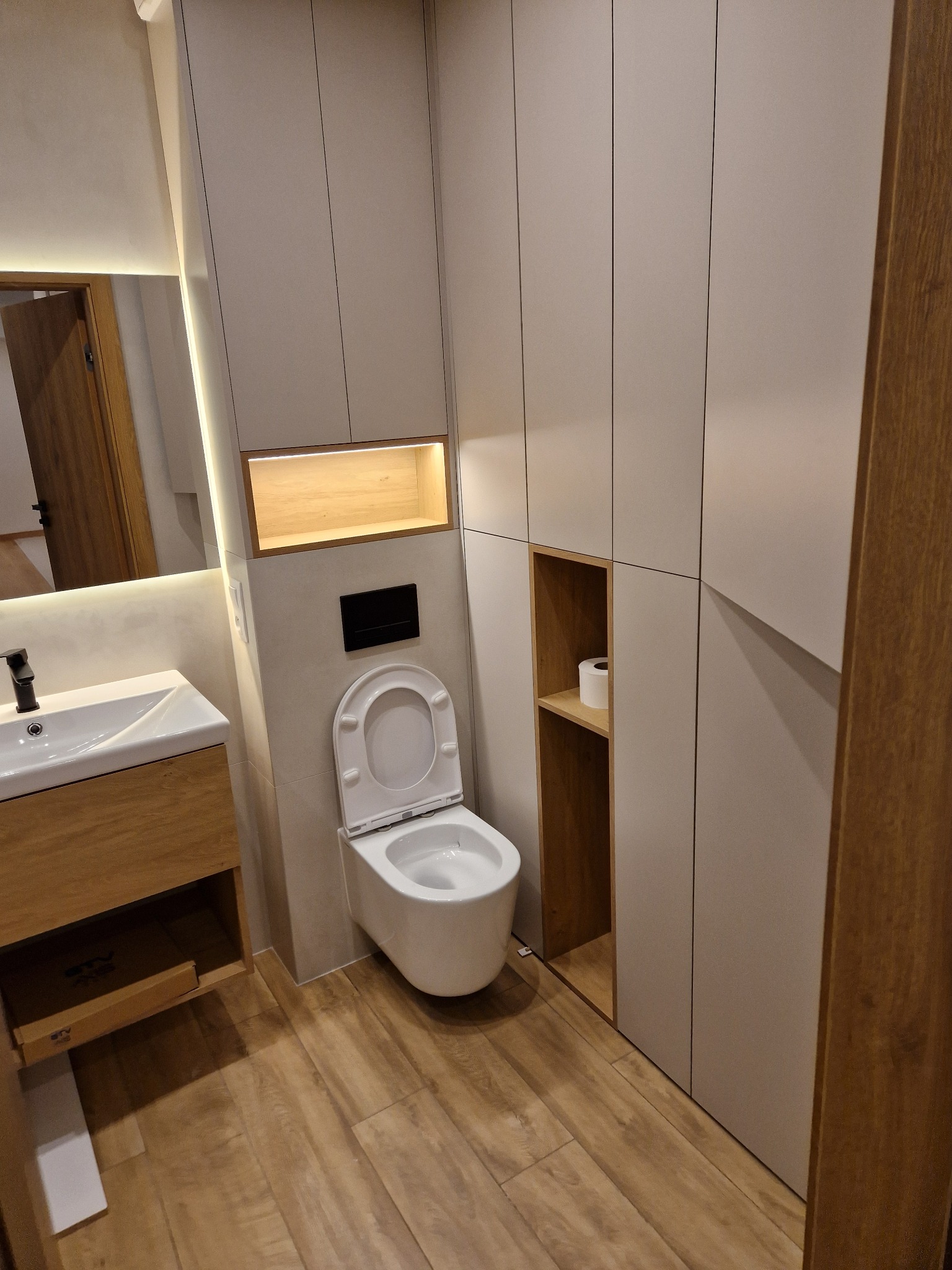 Nowoczesna łazienka po remoncie: podwieszana toaleta, szafki w zabudowie, drewniana podłoga i umywalka z lustrem. Minimalistyczny design z oświetleniem LED.