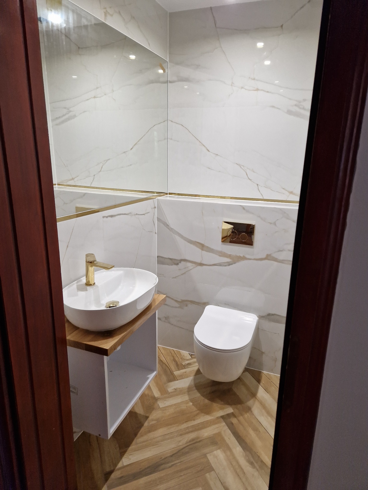 Elegancka łazienka z umywalką nablatową i złotą armaturą, wykończona płytkami imitującymi marmur i drewnianą podłogą ułożoną w jodełkę, widziana zza uchylonych drzwi.