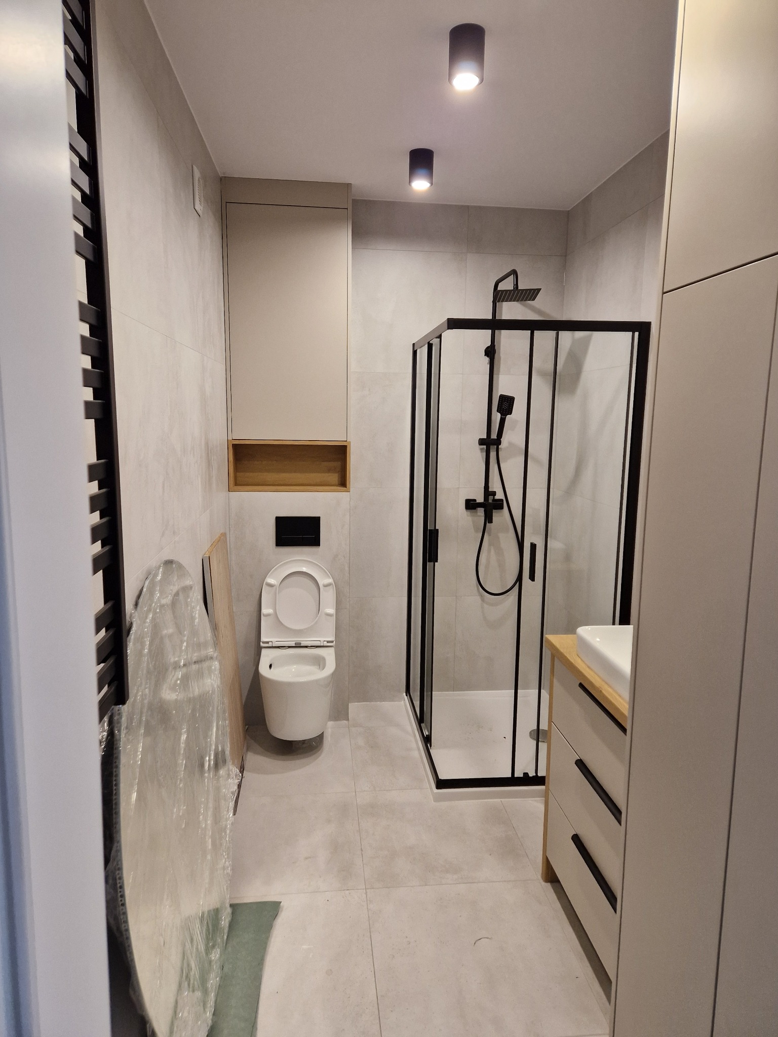 Nowoczesna łazienka po remoncie: kabina prysznicowa z czarną armaturą, toaleta podwieszana, szara glazura i szafka z umywalką. Widoczne elementy w trakcie montażu.