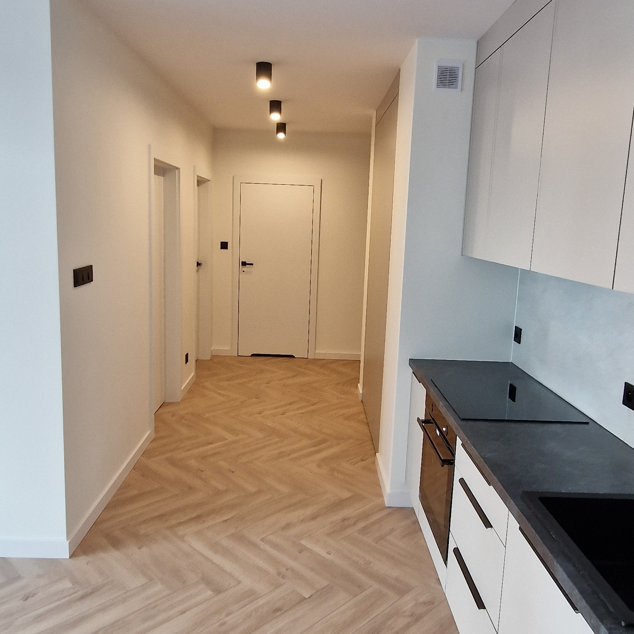 Nowoczesne wnętrze: jasna kuchnia z czarnym blatem i parkietem w jodełkę, widok na korytarz z białymi drzwiami i minimalistycznym oświetleniem.