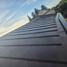 Czas na Mycie - Błyszczący, ciemny dach z blachy na tle błękitnego nieba z chmurami. Widoczny fragment domu i drzewa. Ujęcie z dołu, podkreślające geometrię dachu.