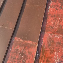 Czas na Mycie - Częściowo odnowiony, brązowy dach z blachy. Widoczne fragmenty starej, czerwonej farby i narzędzie do malowania. Ujęcie z góry, w słonecznym świetle.