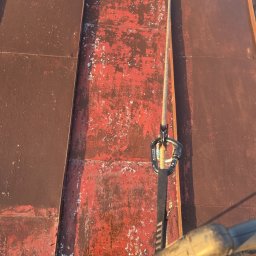 Czas na Mycie - Czerwono-brązowy dach z blachy z widocznymi śladami korozji i łuszczącej się farby. Lina asekuracyjna z karabińczykiem. Widok z góry, podkreślający fakturę i stan dachu.