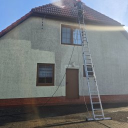 Czas na Mycie - Pracownik na drabinie czyści elewację domu strumieniem wody pod ciśnieniem. Widoczny fragment umytej ściany, ceglana podmurówka i dach z czerwonej dachówki.