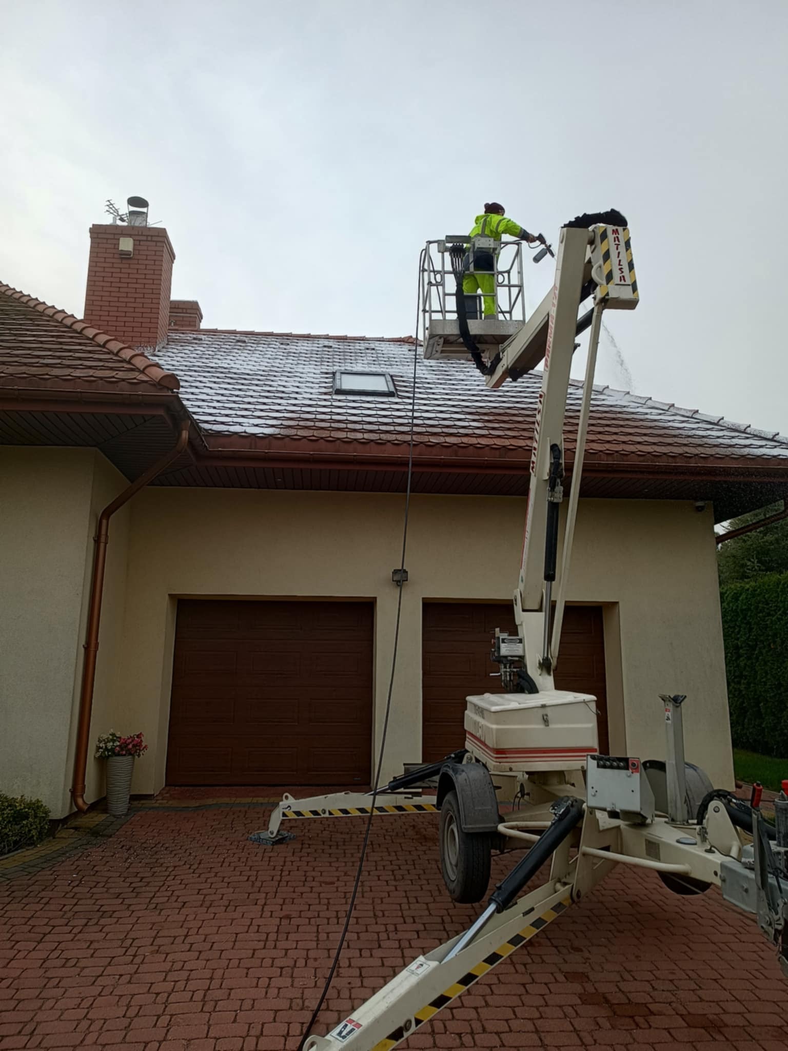 Pracownik w żółtym kombinezonie na podnośniku koszowym czyści dach domu jednorodzinnego strumieniem wody pod ciśnieniem. Widoczny ceglany komin i brązowe rynny.