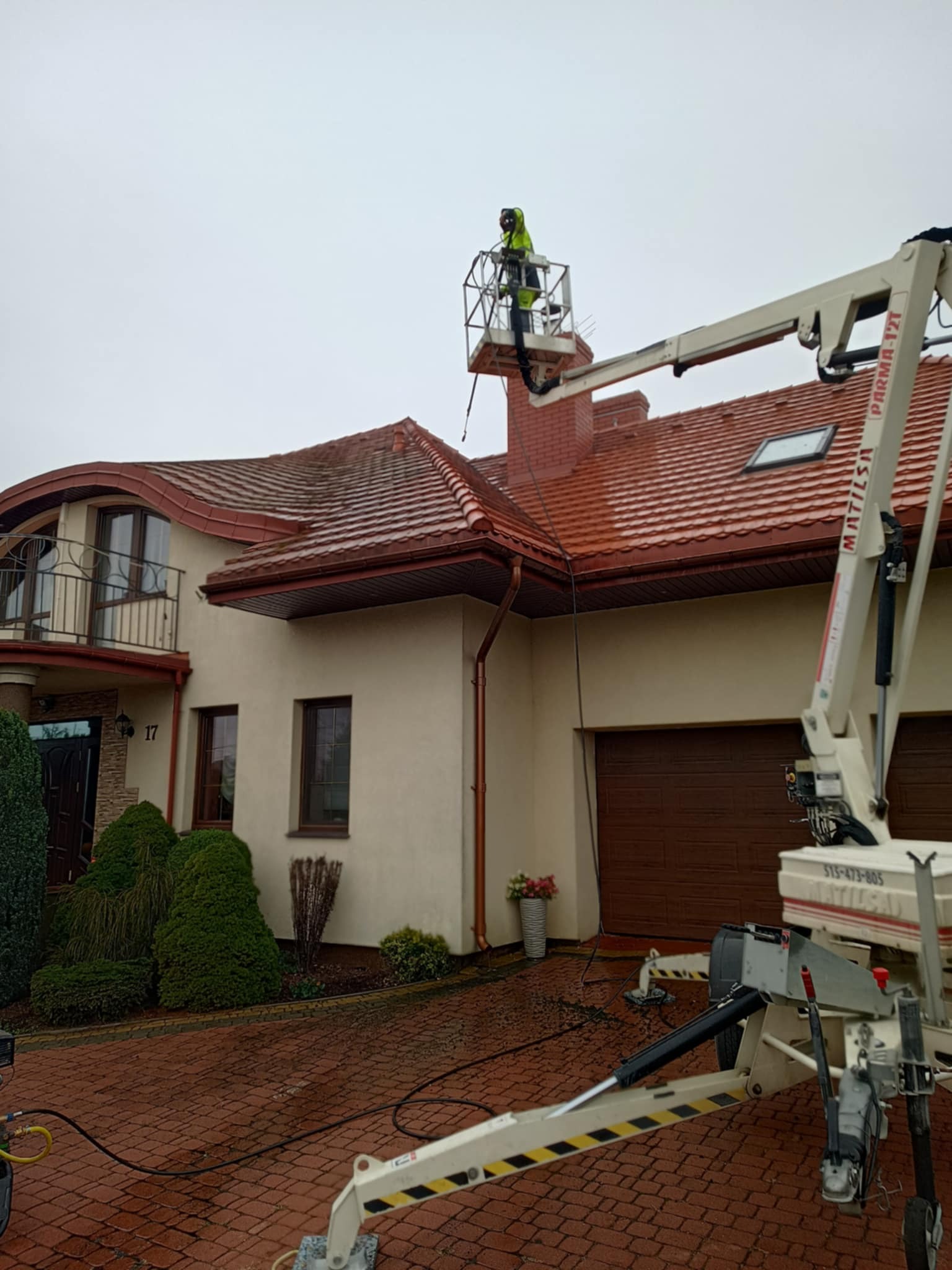 Pracownik na podnośniku czyści dach z czerwonej dachówki w Łapach. Widoczny fragment elewacji domu i podjazd z kostki. Prace na wysokości.