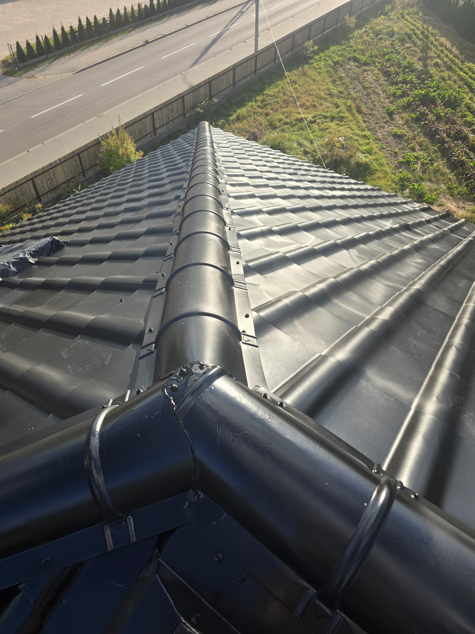 Błyszczący, czarny dach z dachówki ceramicznej, z widocznymi elementami orynnowania. Ujęcie z góry, w tle zieleń i droga.