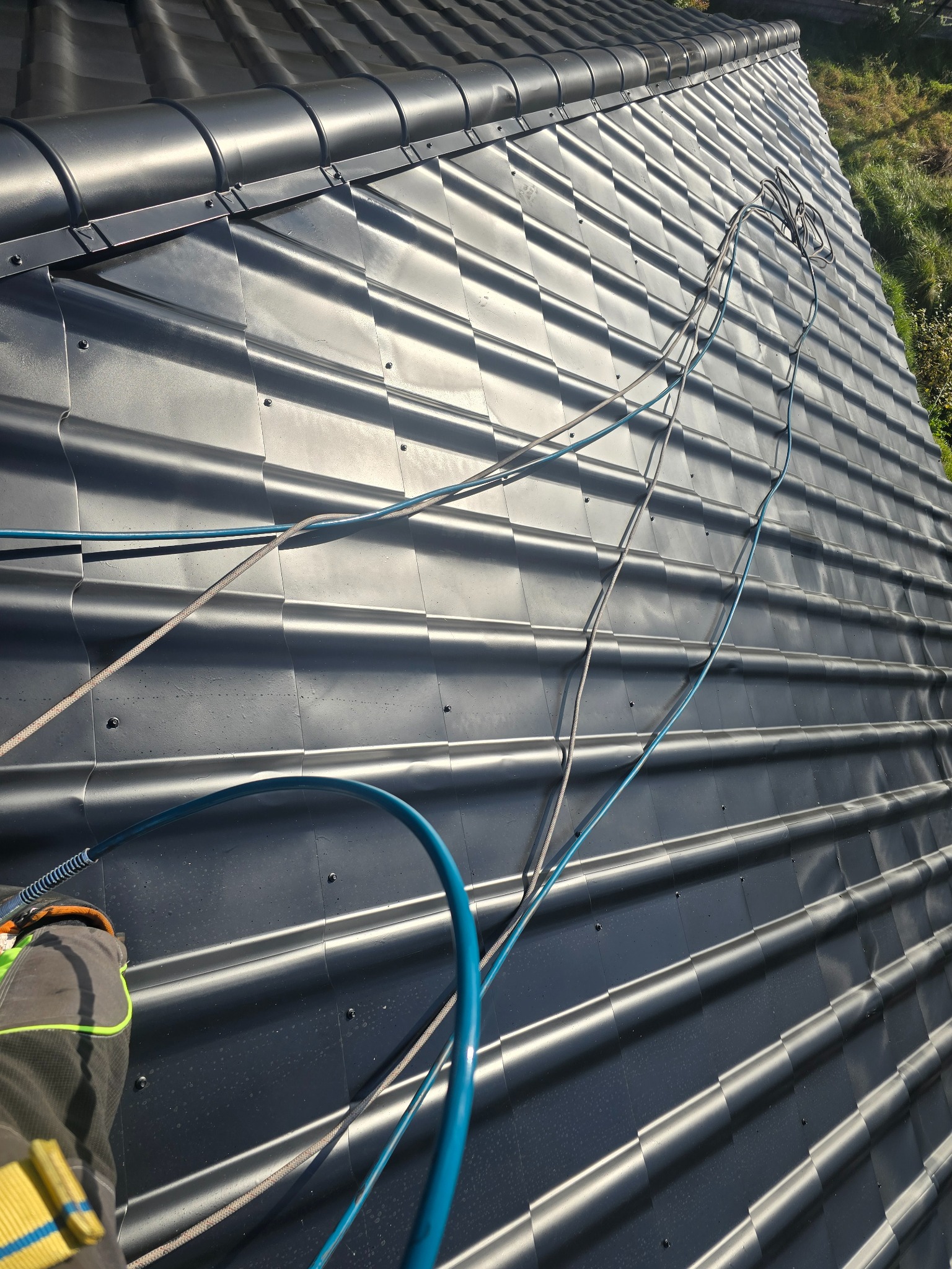 Błyszczący, grafitowy dach z blachy trapezowej, zabezpieczony linami asekuracyjnymi. Widoczny fragment kombinezonu roboczego. Ujęcie z góry, w słoneczny dzień.