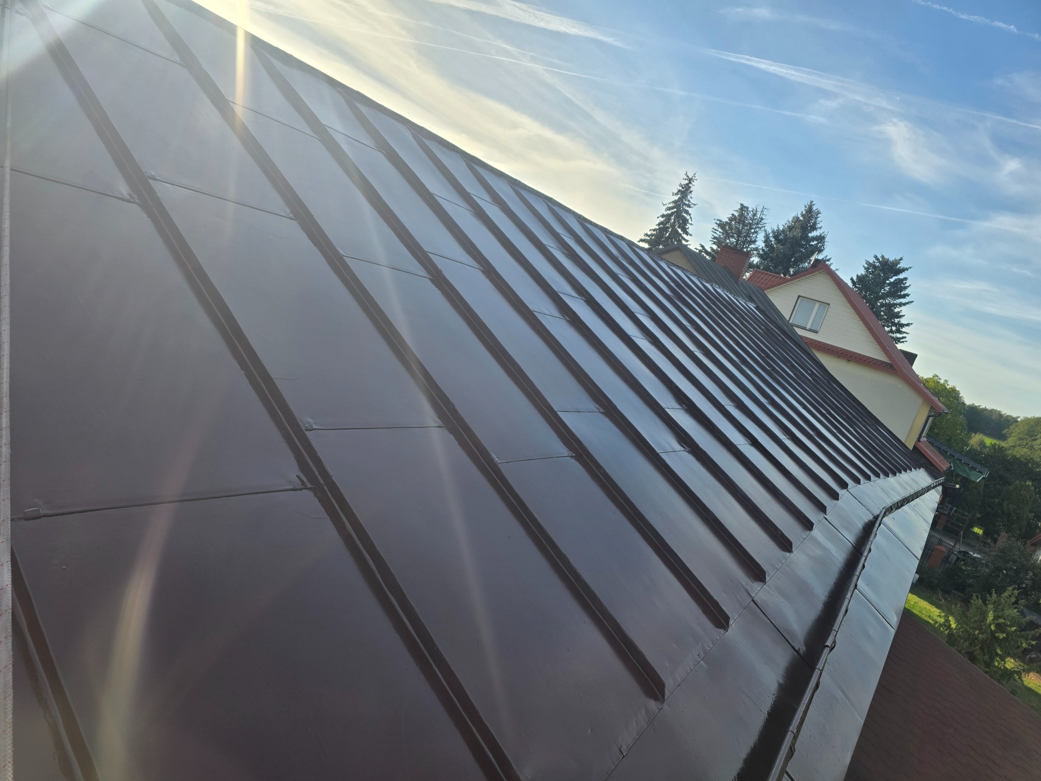Błyszczący, brązowy dach z blachy trapezowej w promieniach słońca, widok z dołu, fragment budynku w tle, błękitne niebo z chmurami.