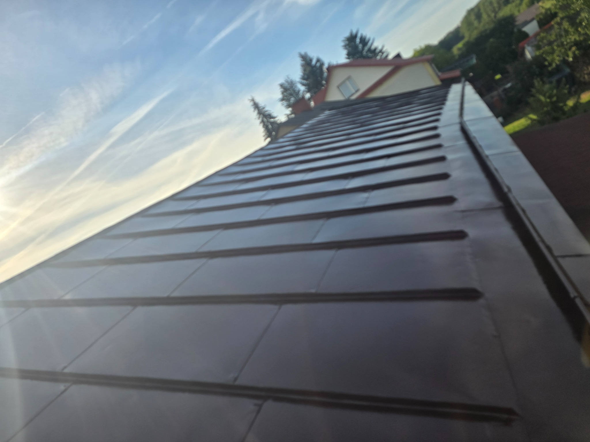 Błyszczący, ciemny dach z blachy na tle błękitnego nieba z chmurami. Widoczny fragment domu i drzewa. Ujęcie z dołu, podkreślające geometrię dachu.