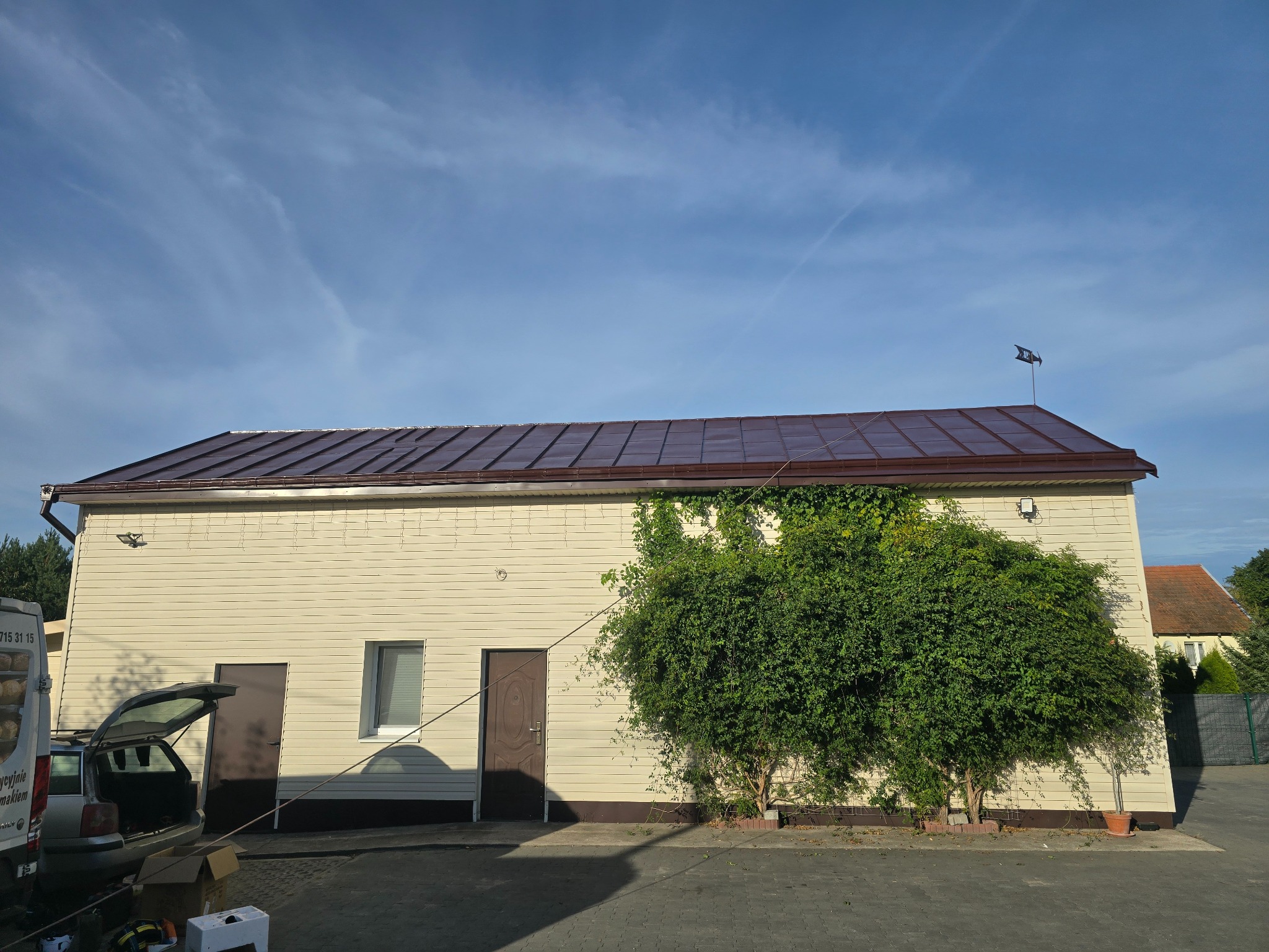 Budynek z brązowym dachem i beżową elewacją, częściowo porośnięty zielenią. Widoczny fragment samochodu dostawczego i akcesoria na ziemi. Błękitne niebo z delikatnymi chmurami.