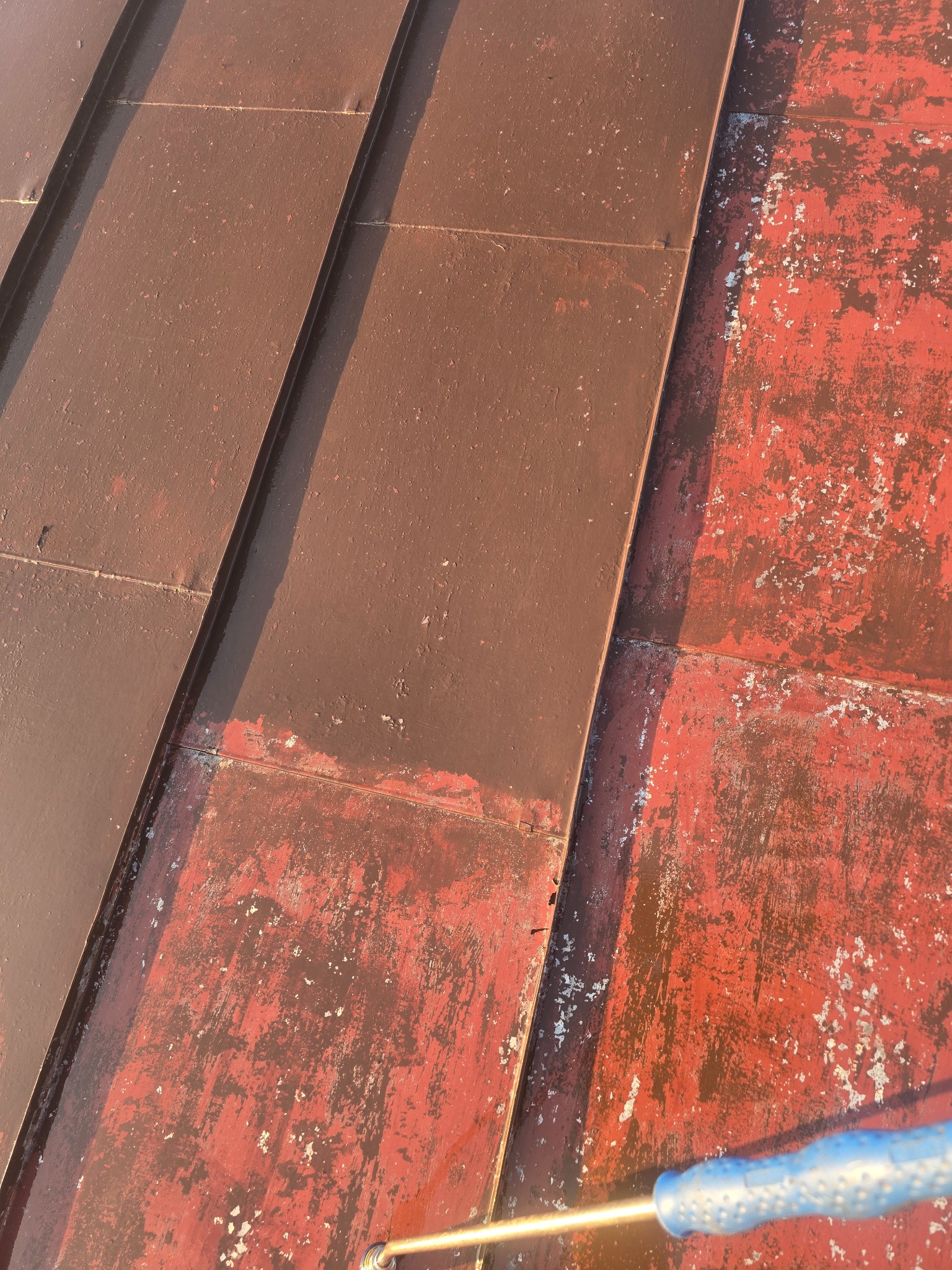 Częściowo odnowiony, brązowy dach z blachy. Widoczne fragmenty starej, czerwonej farby i narzędzie do malowania. Ujęcie z góry, w słonecznym świetle.