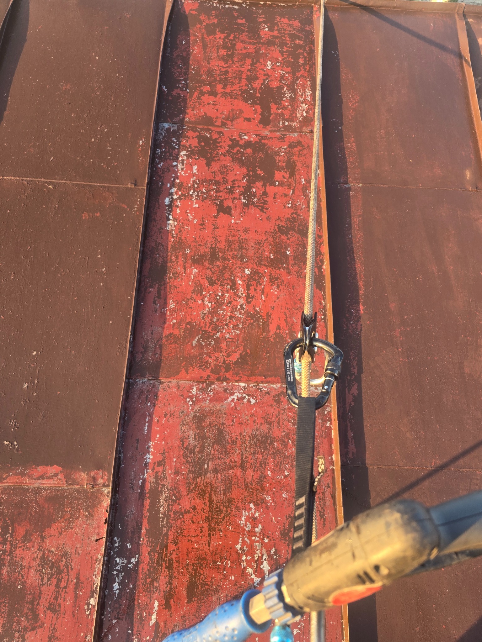 Czerwono-brązowy dach z blachy z widocznymi śladami korozji i łuszczącej się farby. Lina asekuracyjna z karabińczykiem. Widok z góry, podkreślający fakturę i stan dachu.