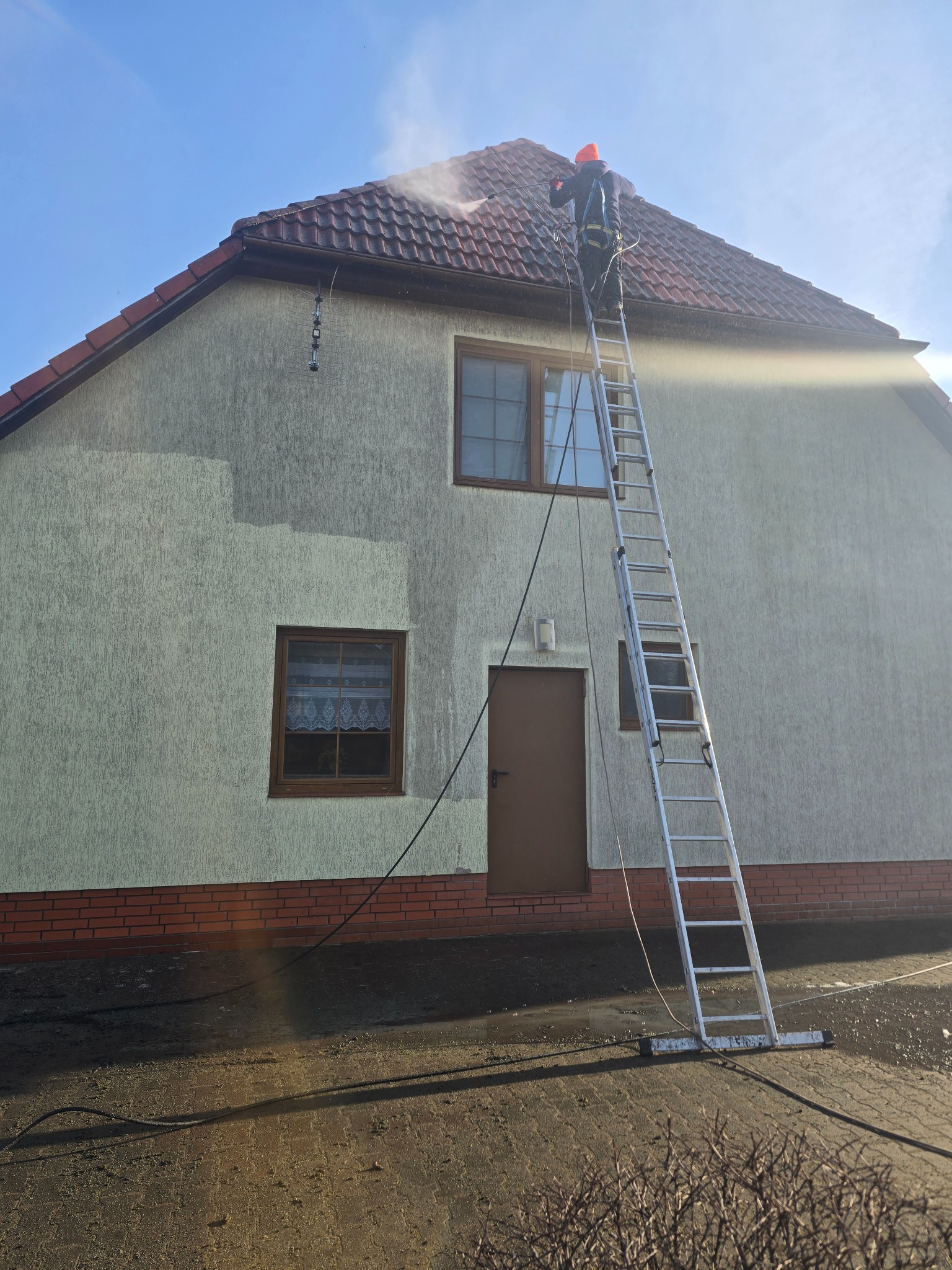 Pracownik na drabinie czyści elewację domu strumieniem wody pod ciśnieniem. Widoczny fragment umytej ściany, ceglana podmurówka i dach z czerwonej dachówki.
