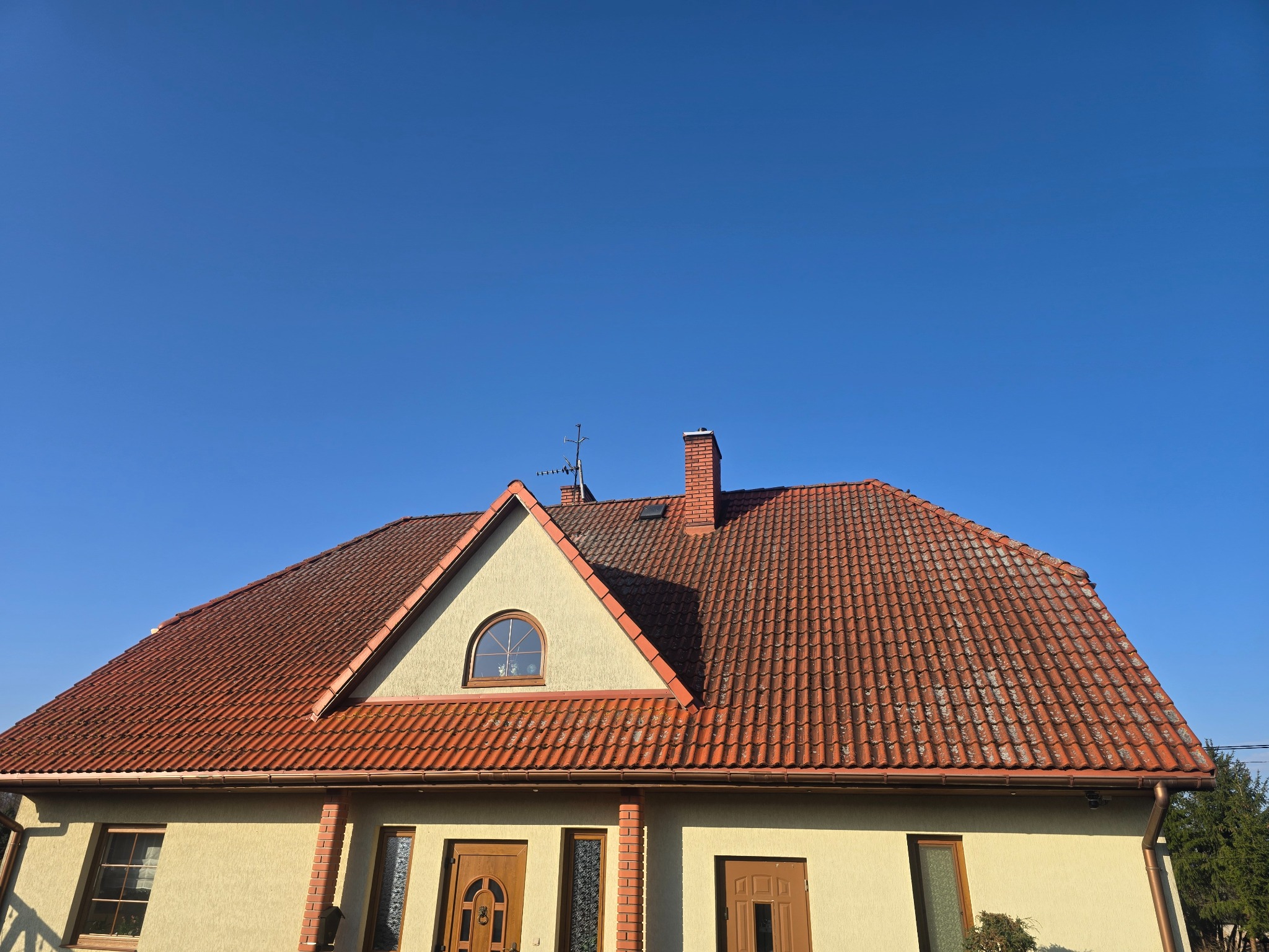 Dom jednorodzinny z czerwoną dachówką i beżową elewacją, widoczny z dołu na tle błękitnego nieba. Charakterystyczne okrągłe okno na poddaszu.