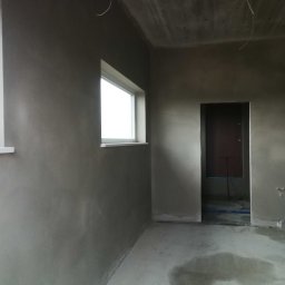 Patryk Bredka - Surowe wnętrze po tynkowaniu ścian i wylewce podłogi, widoczne okna i drzwi, przygotowane do dalszych prac wykończeniowych. Minimalistyczny kadr, surowy beton, naturalne światło.