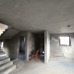 Patryk Bredka - Surowe wnętrze domu po tynkowaniu: schody betonowe, ściany pokryte szarym tynkiem, widoczne okna i krzesło. Wnętrze w trakcie wykańczania, przygotowane do dalszych prac.