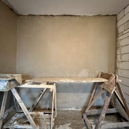Patryk Bredka - Świeżo otynkowana ściana w pomieszczeniu, widoczne drewniane rusztowania i narzędzia murarskie. Fragment ściany z cegły. Szare tynki na ścianach i suficie.