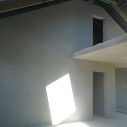 Patryk Bredka - Świeżo otynkowane wnętrze poddasza z widoczną konstrukcją dachu i wpadającym światłem słonecznym, ukazujące gładkie, szare ściany przed dalszym wykończeniem.