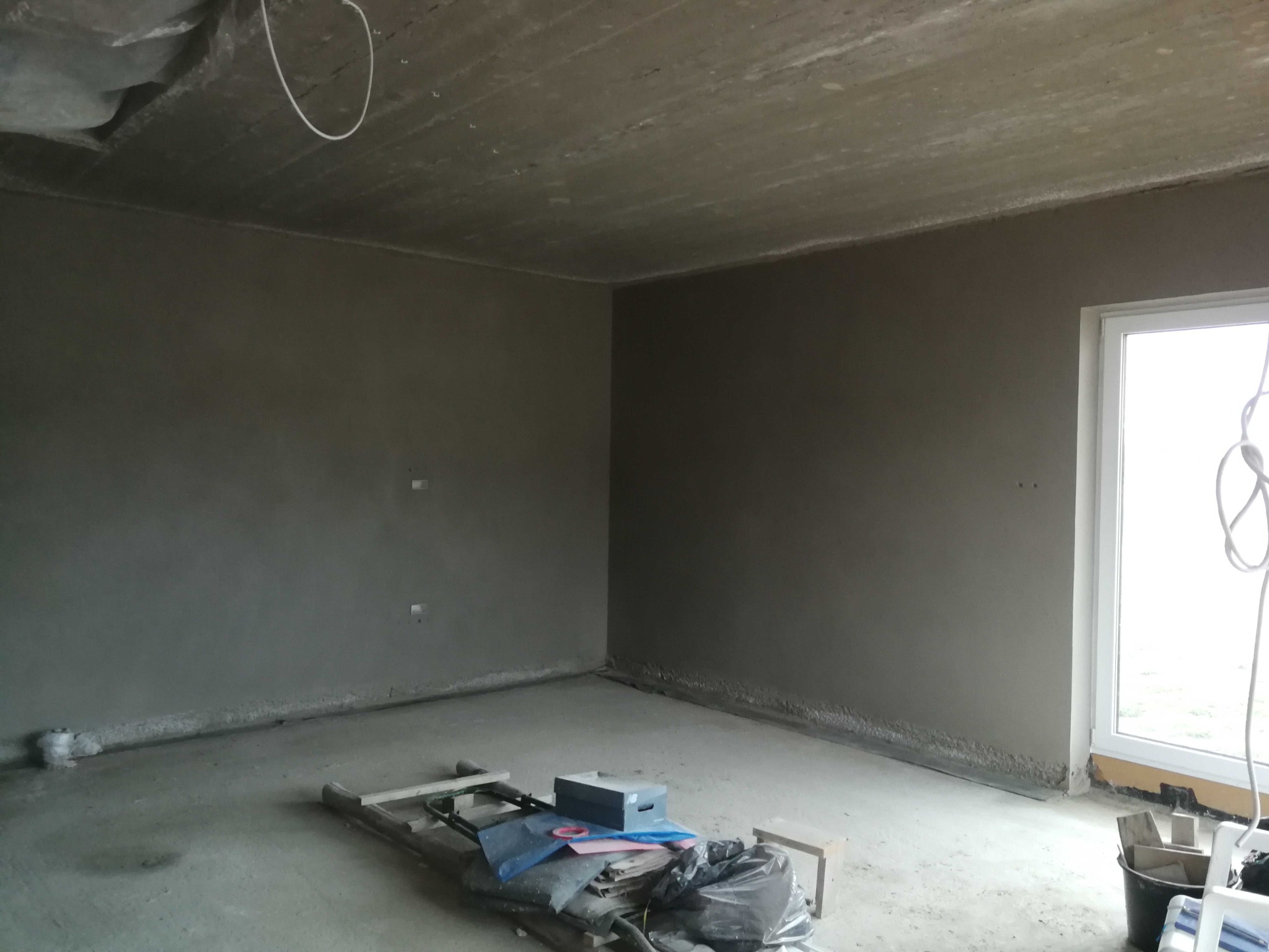 Surowe wnętrze po tynkowaniu ścian, widoczne narzędzia i materiały budowlane, okno z widokiem na zewnątrz, betonowy sufit z wystającymi kablami.