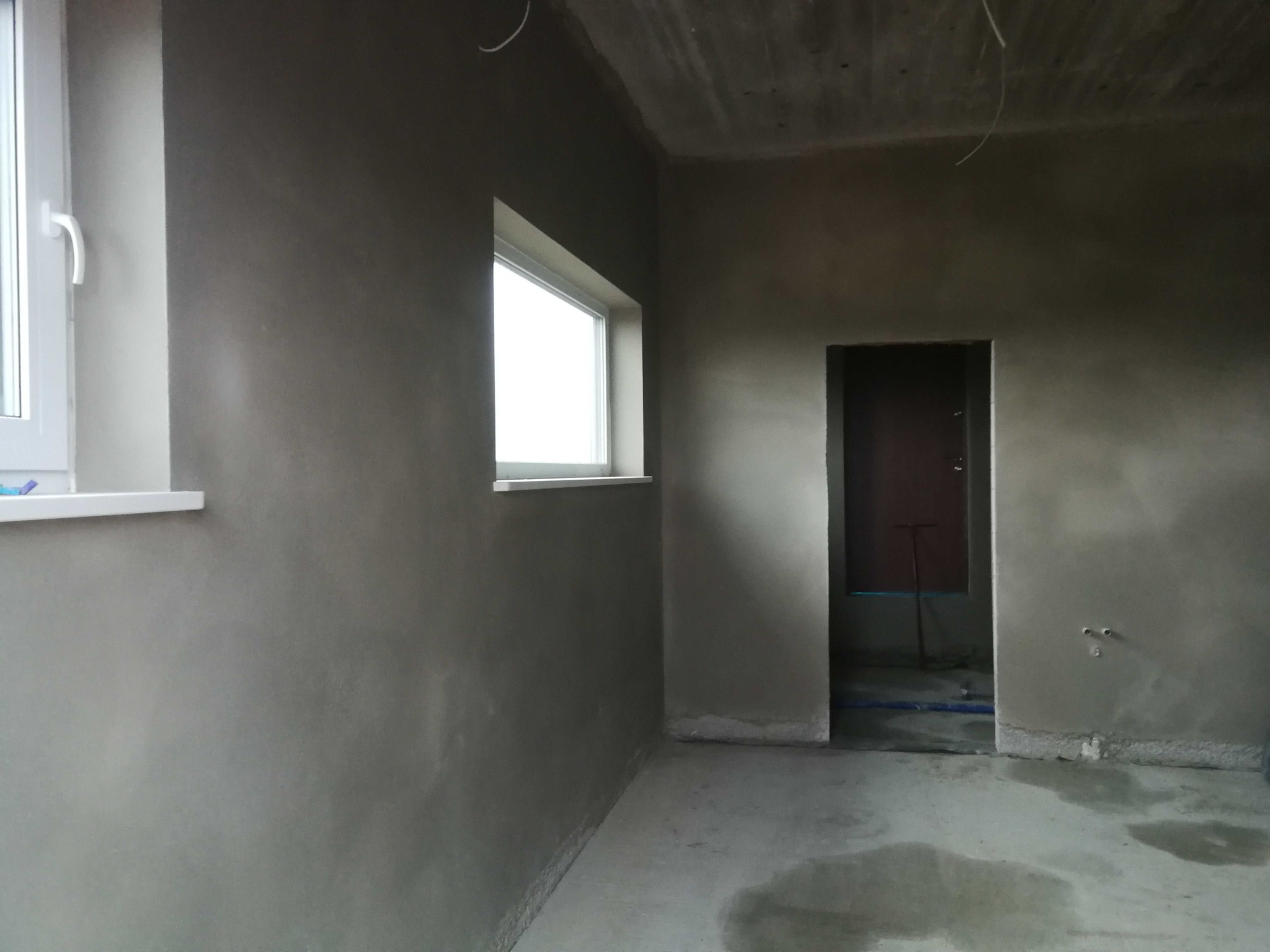 Surowe wnętrze po tynkowaniu ścian i wylewce podłogi, widoczne okna i drzwi, przygotowane do dalszych prac wykończeniowych. Minimalistyczny kadr, surowy beton, naturalne światło.
