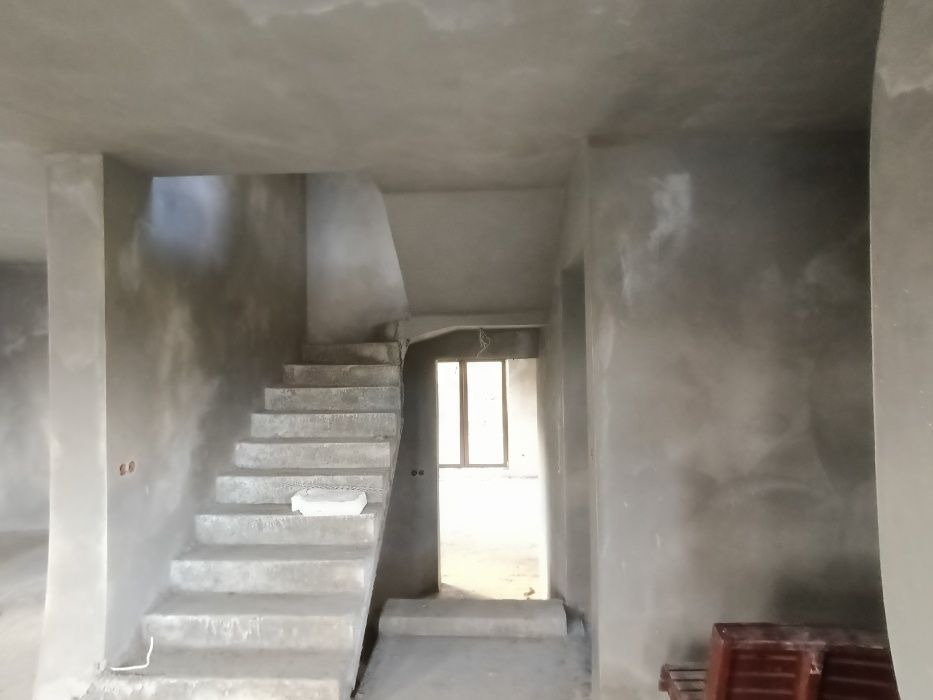 Surowe wnętrze w trakcie tynkowania: betonowe schody, ściany pokryte szarym tynkiem, widoczne niedoskonałości i surowość materiałów. W tle okno z widokiem na zewnątrz.