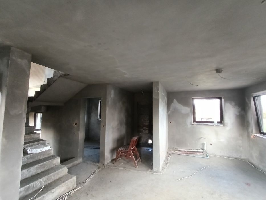 Surowe wnętrze domu po tynkowaniu: schody betonowe, ściany pokryte szarym tynkiem, widoczne okna i krzesło. Wnętrze w trakcie wykańczania, przygotowane do dalszych prac.
